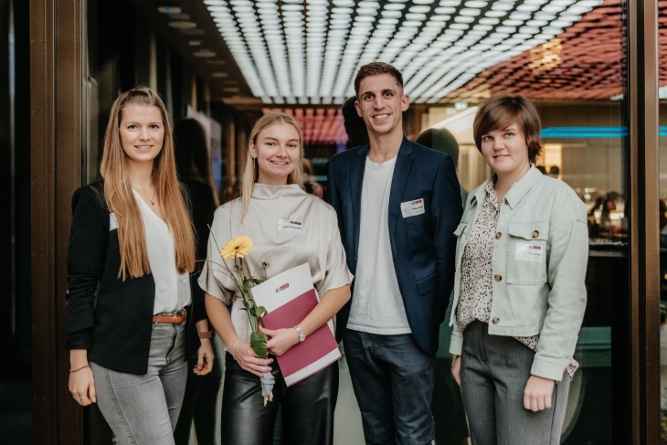 Vier Personen lächeln in die Kamera. Die Personen tragen einen Business Casual Look. Die zweite Person von Links ist die Stipendiatin. Sie hält eine gelbe Blume und eine Zeugnismappe in der Hand. 