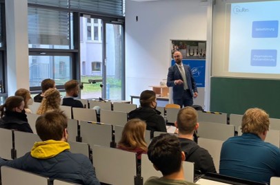 Einblick in die Insidestory. Menschen sitzen in einem Hörsaal und schauen einen Redner an, welcher vorne steht und etwas erzählt.